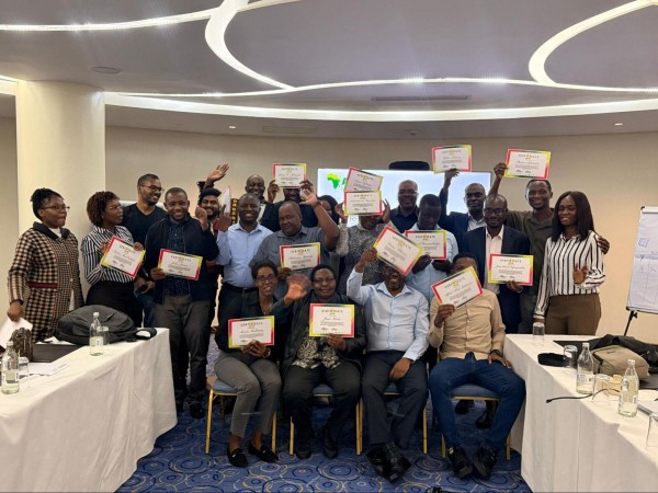 A group of about 20 smiling professionals pose in a modern conference room, holding up "Certificate of Participation" awards. They are arranged in rows behind white-clothed tables, appearing joyful and celebratory. In the background, a screen displays a green logo of the African continent.