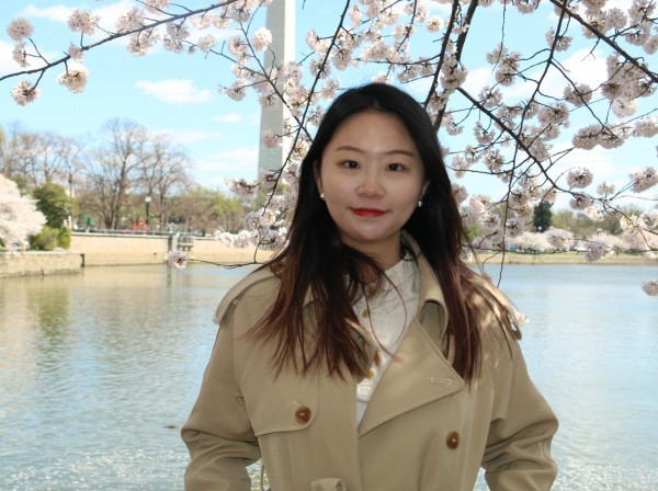 A medium close-up color photograph taken on a sunny day shows an East Asian woman with dark hair in a beige trench coat smiling under cherry blossom branches in Washington, DC. Behind her is the Tidal Basin with the Washington Monument and more cherry trees on the opposite shore under a partly cloudy sky. The lighting is sunny and the photo is taken at eye level.