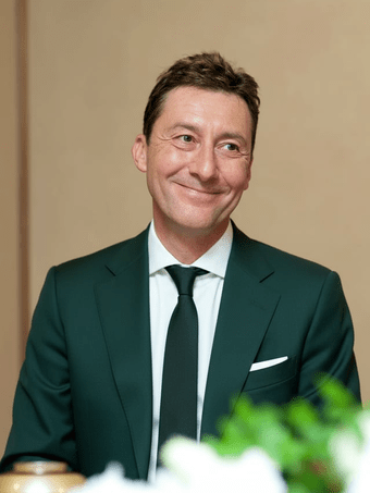 A medium shot of a man with short brown hair and a slight smile, wearing a dark green suit, white shirt, and dark tie against a neutral background.