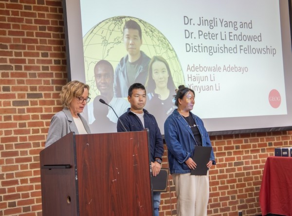 A woman with blonde hair speaks at a podium during an awards ceremony. Behind her, a presentation screen announces the "Dr. Jingli Yang and Dr. Peter Li Endowed Distinguished Fellowship," listing recipients Adebowale Adebayo, Haijun Li, and Xinyuan Li. Two of the honorees stand to the right of the podium against a backdrop of a brick wall.