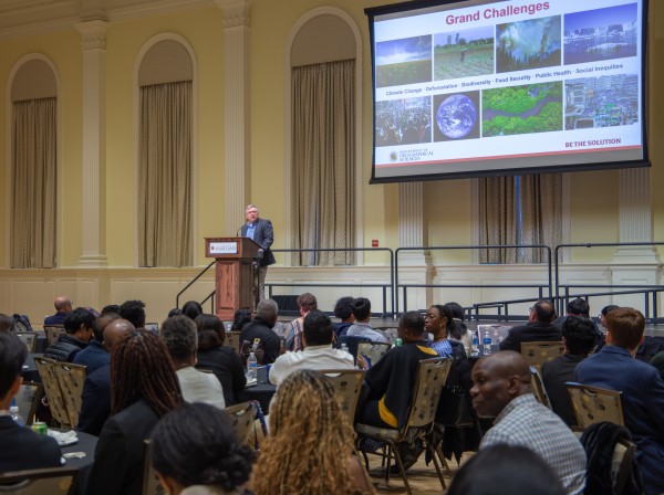 A wide shot from the back of a large, high-ceilinged ballroom where an audience is seated at round tables, facing a speaker at a wooden lectern on a stage. The speaker, a man in a dark suit jacket, stands next to a large projection screen displaying a presentation titled "Grand Challenges." The slide features a collage of environmental and social images with captions like "Climate Change," "Deforestation," and "Food Security," alongside the logo for the University of Maryland Department of Geographical Scie