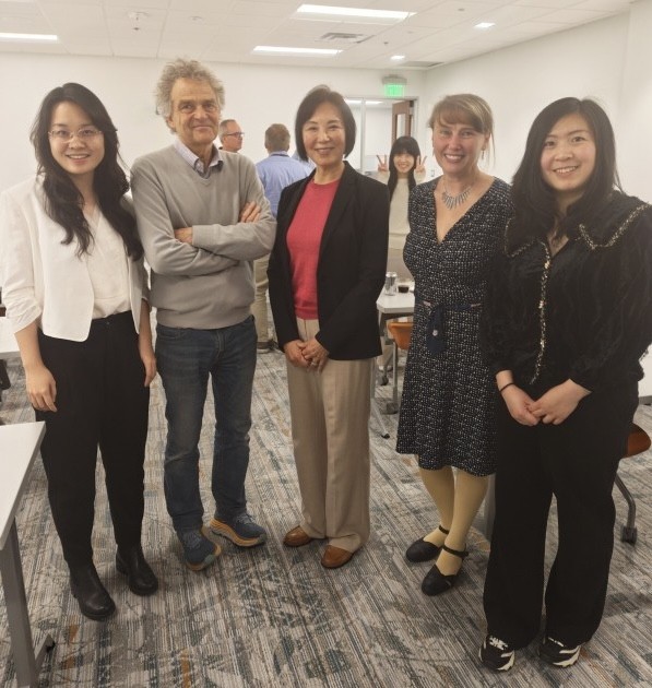 A group of five people pose in a bright, modern room. From left to right, they are a student in a white blazer, a professor with grey hair and crossed arms, an distinguished woman in a black blazer and pink top, another professor in a patterned dress, and a student in a black textured top. In the background, a woman playfully peeks out while making peace signs.