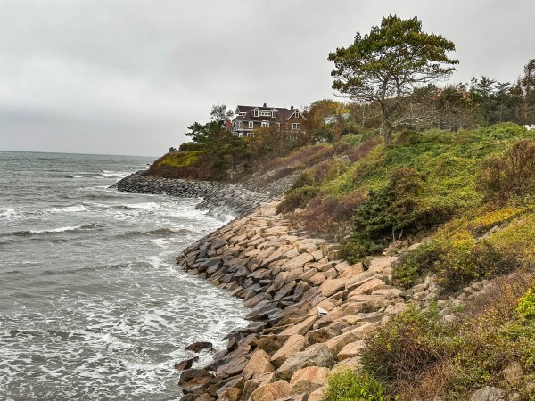 A large, multi-story coastal house sits atop a grassy bluff overlooking a rocky shoreline. Under a gray, overcast sky, small waves from the ocean crash against a heavy stone revetment that lines the coast to prevent erosion. The hillside is covered in green and autumnal shrubbery, with a single tall, bare-branched tree standing in the foreground.