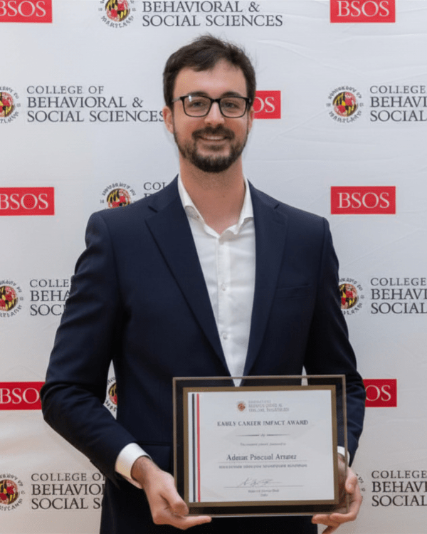 A smiling man with glasses and a beard, wearing a dark suit jacket and white shirt, stands in front of a University of Maryland backdrop while holding an "Early Career Impact Award" certificate.