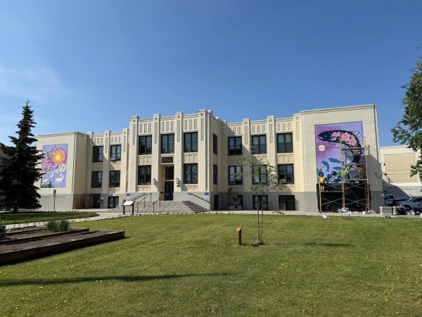 A light-beige Art Deco city hall building under a clear blue sky, featuring two large, colorful nature-themed murals on its outer wings. The left mural shows mountains and caribou, while artists on scaffolding work on the right mural, which depicts a large fish and wild berries.