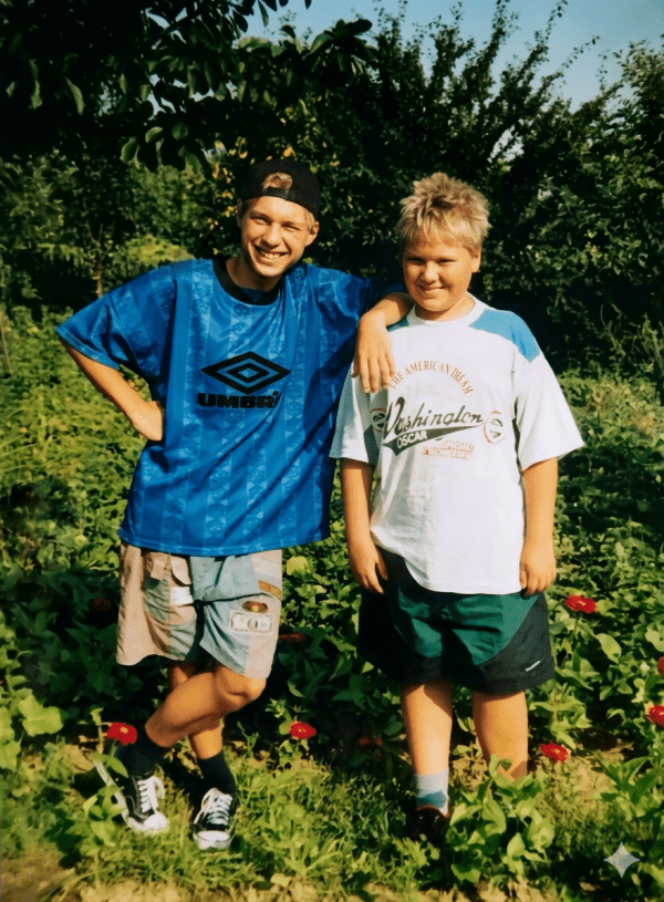 An outdoor photograph of Sergii Sakun standing with his arm around his brother in a sunny garden. On the left, Sergii wears a backward black cap, a large blue Umbro jersey, and patchwork shorts. On the right, his younger brother wears a white graphic t-shirt and green shorts. Both are smiling warmly against a background of lush green bushes and red flowers.