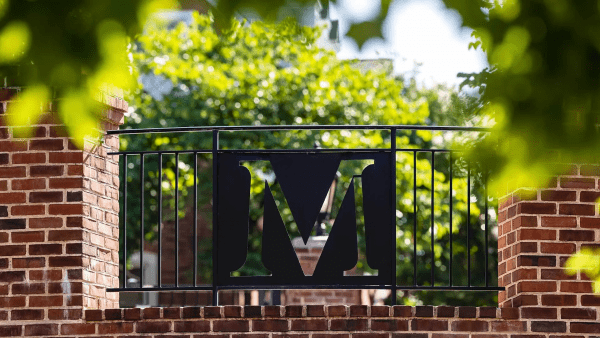 A close-up shot of a black metal gate panel featuring a large, stylized letter "M" cutout. The panel is set within a black metal railing between two brick pillars, framed by blurry green tree leaves in the foreground and background.