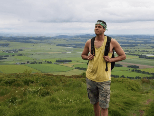 A man with a backpack and a green bandana stands on a grassy hilltop, looking off to the side with a vast landscape of green fields stretching out behind him under an overcast sky.