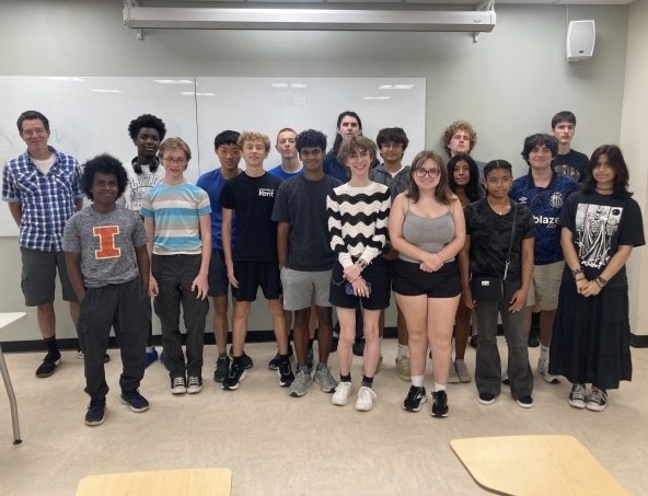 Associate Research Professor Evan Ellicott stands on the far left next to a group of high school students in a classroom. On the screen behind them, a slide titled "Satellite Image Analysis Project" displays satellite imagery and student presentation details.