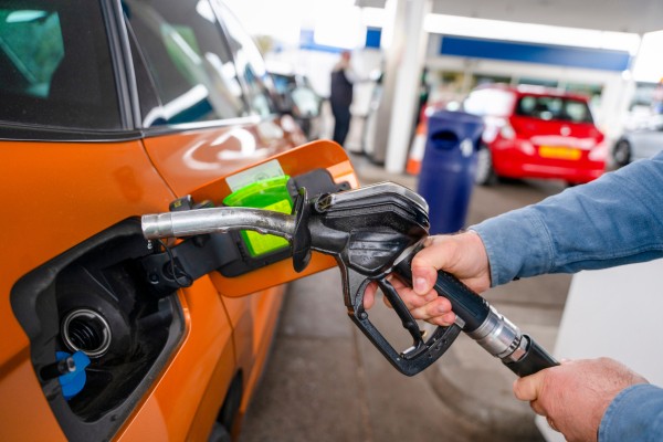Close-up of a person's hands holding a black fuel nozzle and removing it from the open fuel tank door of an orange car at a gas station.