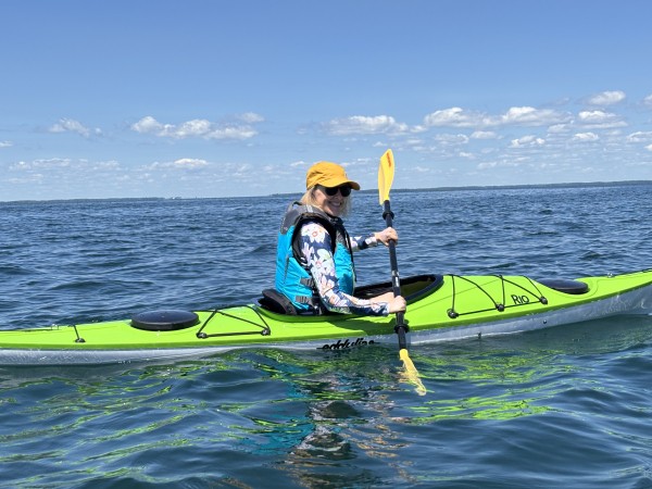 A person smiling while paddling a bright lime green kayak on calm, open blue water under a clear sunny sky.