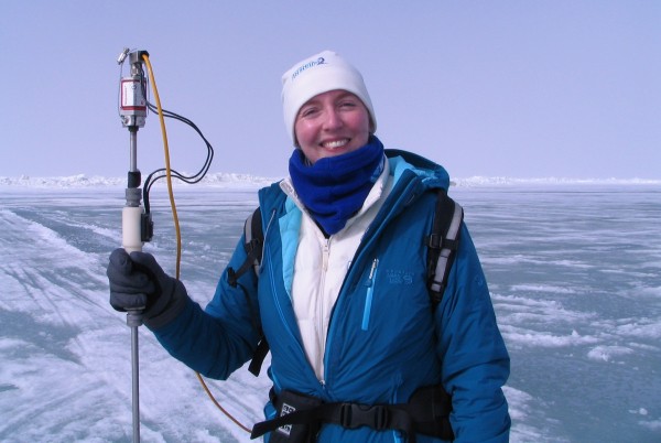 A smiling researcher wearing cold-weather gear—including a blue jacket, white beanie, and blue neck gaiter—stands on a vast, frozen icy landscape. She is holding a vertical scientific probe or measurement device attached to a yellow cable under a clear, bright sky.
