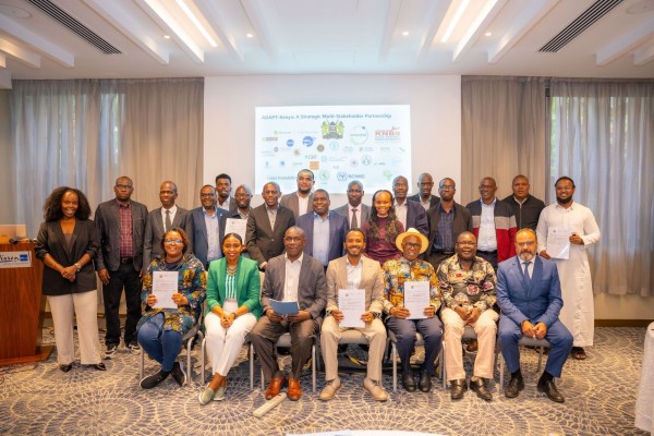 A group of over twenty business professionals posing for a group photo in a conference room. Seven people are seated in chairs in the front row—several holding signed documents or folders—while a larger group stands behind them. In the background, a presentation screen displays the title "ADAPT-Kenya: A Strategic Multi-Stakeholder Partnership" along with various organization logos.