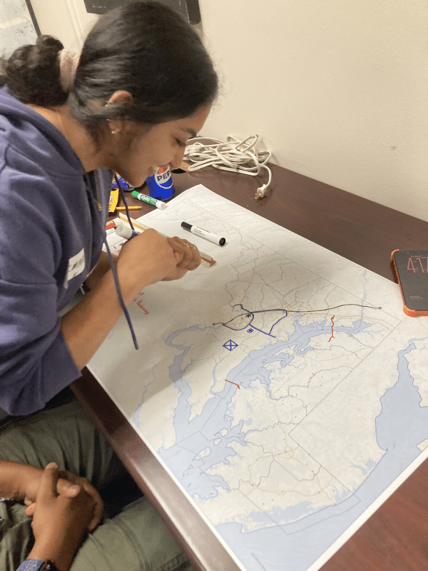 A person leaning over a wooden desk, using a scale ruler and marker to draw routes and symbols on a large paper map laid out in front of them.   