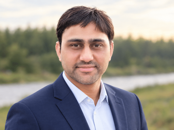 Headshot of Abdul Qadir smiling in a navy blue suit blazer and light blue dress shirt, outdoors with a soft-focus natural background.