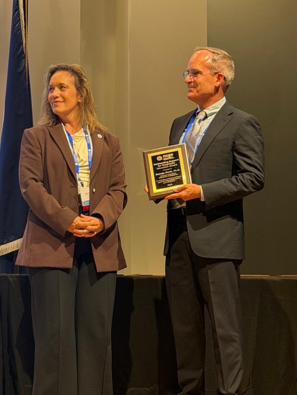 Professor George Hurtt in a suit and glasses holding an NCGE "Outstanding Support for Geography Education Award" plaque next to a smiling Celeste Reynolds  in a brown blazer, standing on stage near a flag.   