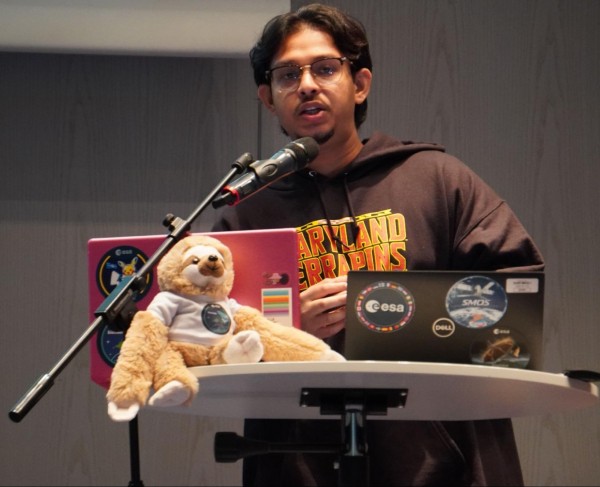 A young man wearing glasses and a black "Maryland Terrapins" hoodie speaking into a microphone at a podium. In front of him sits a small plush sloth wearing a white t-shirt next to a pink laptop and a black Dell laptop covered in ESA (European Space Agency) and space-themed stickers.