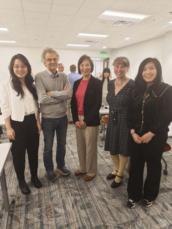 A group of five people pose in a bright, modern room. From left to right, they are a student in a white blazer, a professor with grey hair and crossed arms, an distinguished woman in a black blazer and pink top, another professor in a patterned dress, and a student in a black textured top. In the background, a woman playfully peeks out while making peace signs.