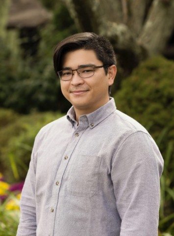 A young man in glasses and a long-sleeved button-down shirt poses in front of a flowering garden.