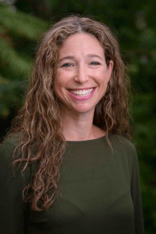 A woman with long, wavy hair smiles at the camera agains a green forest background.