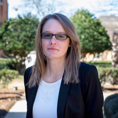  A woman in glasses and a blazer stands outside, smiling confidently against a blurred urban park background.