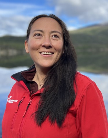 This is a portrait of a smiling woman with long dark hair, wearing a bright red jacket against a vivid natural background of a calm lake and distant, snow-capped mountains.