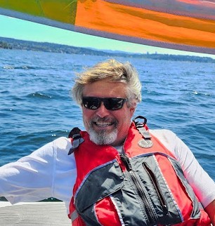 A smiling man wearing a life jacket and sunglasses sits on a boat, enjoying a sunny day on the water.