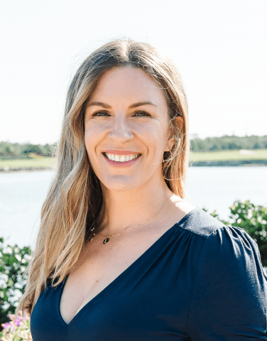 A light-skinned woman with blonde hair is smiling in front of a like.