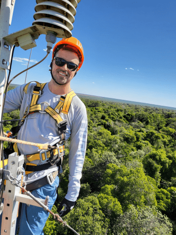 Man wearing a helmet, sunglasses and climbing gear is at the top of a telecommunications tower overseeing a forest canopy..