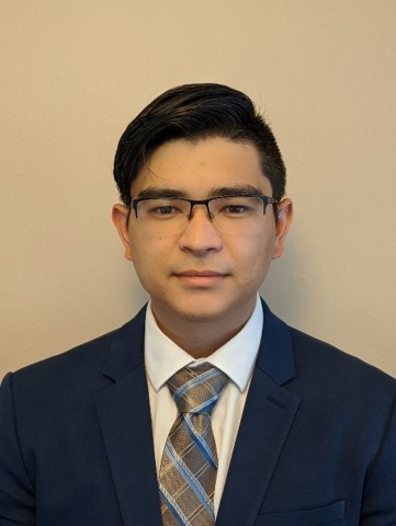 A professional headshot of a young man with dark hair and glasses, wearing a navy blue suit, a white dress shirt, and a blue and tan patterned tie. He is looking directly at the camera with a neutral expression against a plain, light-colored background.