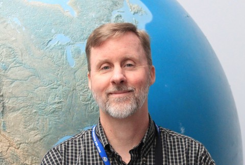 A headshot of a middle-aged man with golden brown hair and a graying beard, smiling in front of a large blue globe.