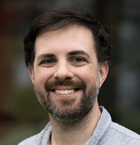 A headshot of a smiling man with dark, slightly messy hair and a short, groomed beard. He is wearing a light gray or blue button-down shirt. The background is softly blurred with hints of green and dark tones.