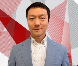 A headshot of a man with short black hair, smiling slightly at the camera. He is wearing a light blue blazer over a white patterned button-down shirt. The background is a modern, abstract geometric design consisting of sharp red, grey, and white triangular shapes with fine diagonal line patterns.
