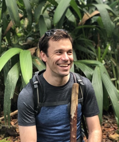 A man with short brown hair smiles while sitting in a lush tropical forest. He is wearing a grey and navy blue t-shirt, a hiking backpack, and sunglasses propped on his head. He holds a tall, unvarnished wooden walking stick in front of him. Thick green palm-like leaves create a dense background.