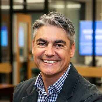 A headshot of a smiling middle-aged man with salt-and-pepper hair, wearing a dark blazer over a blue and white checkered button-down shirt, in a blurred office setting.