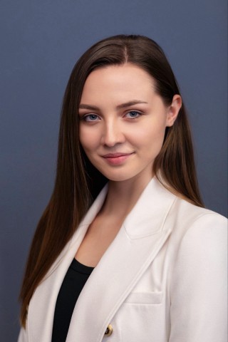 Professional studio headshot of a young woman with long, straight brown hair parted slightly to the side. She has blue eyes, a light complexion, and a soft, confident smile, looking directly at the camera. She is wearing a structured white blazer with a visible lapel and a gold button over a black top. The background is a solid, neutral slate-blue color.