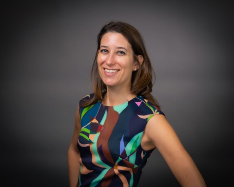 Professional studio headshot of a smiling woman with shoulder-length brown hair, wearing a sleeveless dress with a colorful, abstract geometric pattern, against a solid dark gray background.