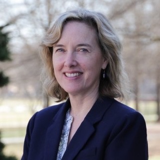 A headshot of a smiling woman with wavy, light blonde or grey hair, looking directly at the camera. She is wearing a dark blue blazer over a light, patterned shirt and small dangle earrings. The background features a softly blurred outdoor setting with trees and daylight.