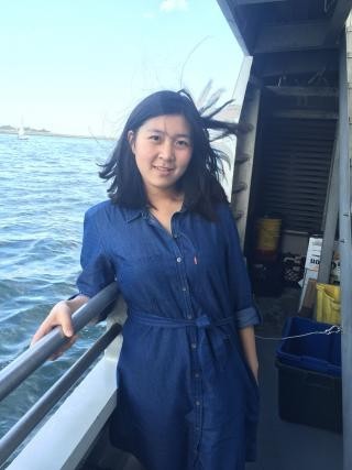 A medium shot of a young woman with shoulder-length dark hair, smiling gently on the deck of a boat. She is wearing a blue denim button-up shirt-dress tied at the waist, and her hair is slightly windswept. She is leaning against a metal railing on the left, looking toward the camera. In the background, there is a body of water under a clear blue sky, with a small sailboat visible in the distance. The right side of the frame shows part of the boat's interior structure and storage items.