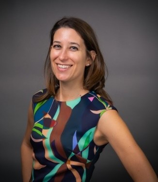 Studio portrait of a smiling woman with shoulder-length brown hair wearing a colorful patterned sleeveless dress against a dark grey background.