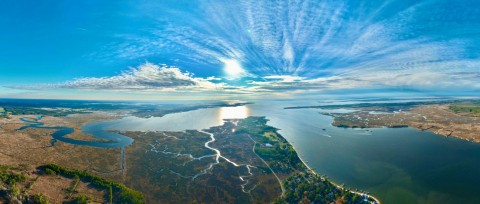 Wide panoramic aerial view of Maryland coastal wetlands, winding tidal waterways, and shoreline trees leading to a vast bay illuminated by a bright sun beneath a wispy blue sky.