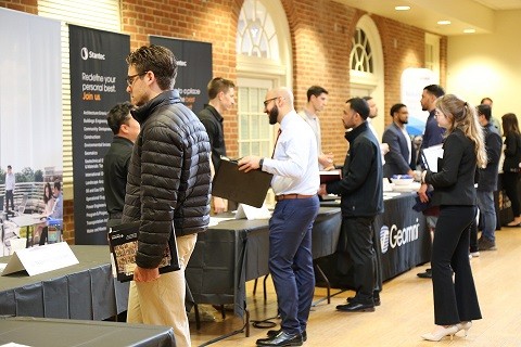 People in professional attire conversing and networking at information tables during a career fair, with company banners like Stantec and Geomni visible in the background.