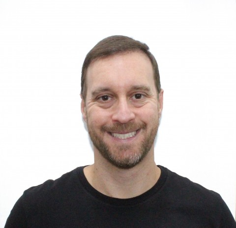 Headshot of a man with short brown hair, brown eyes, and a light beard, smiling at the camera against a plain white background.