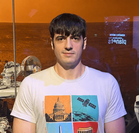 A man with dark hair with bangs wearing a white t-shirt with a four-panel graphic, standing in front of a space exhibit background featuring a Martian landscape and rover equipment.