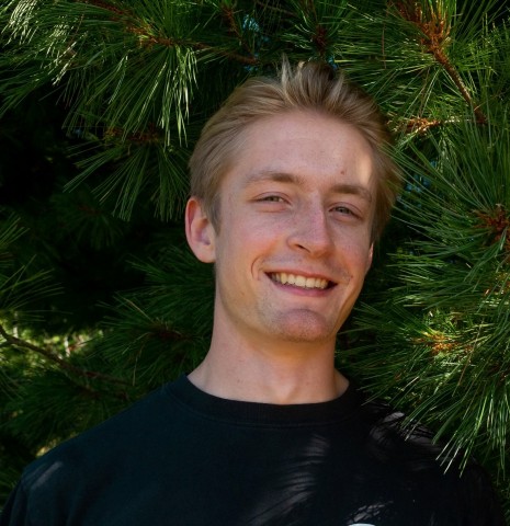 A headshot of a young man with light brown hair smiling, wearing a black t-shirt, set against a background of lush green pine tree branches.   