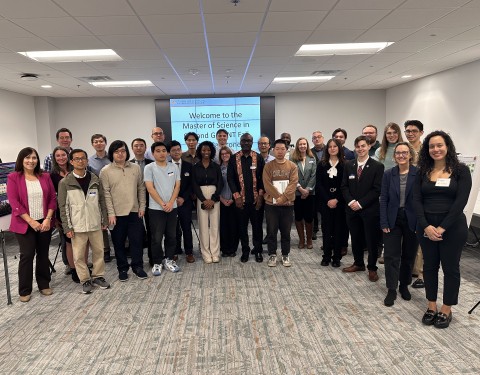 A group of diverse people—students, faculty, and attendees—poses together in a large conference room. They are arranged in multiple rows facing the camera. In the background, a large monitor displays the text: "Welcome to the Master of Science in ... Fall ... Capstone".