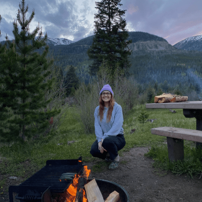A woman enjoying a warm campfire, seated comfortably on the ground, with trees in the background.