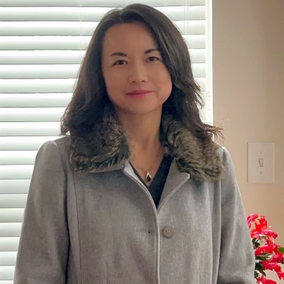 A woman with shoulder-length wavy dark hair smiling warmly at the camera. She is wearing a grey wool coat with a faux-fur collar and a small pendant necklace. She is standing indoors in front of a window with closed white blinds; a red plant is partially visible in the background.