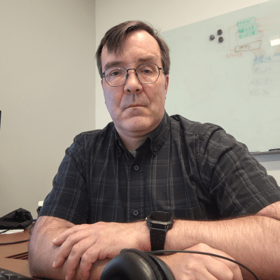 Close-up portrait of a man with short brown hair and round glasses, wearing a dark plaid button-up shirt and a black smartwatch. He is sitting at a wooden desk with his arms crossed, looking forward with a neutral expression.