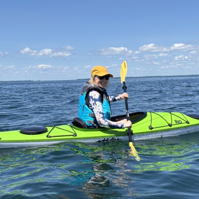 A person smiling while paddling a bright lime green kayak on calm, open blue water under a clear sunny sky.